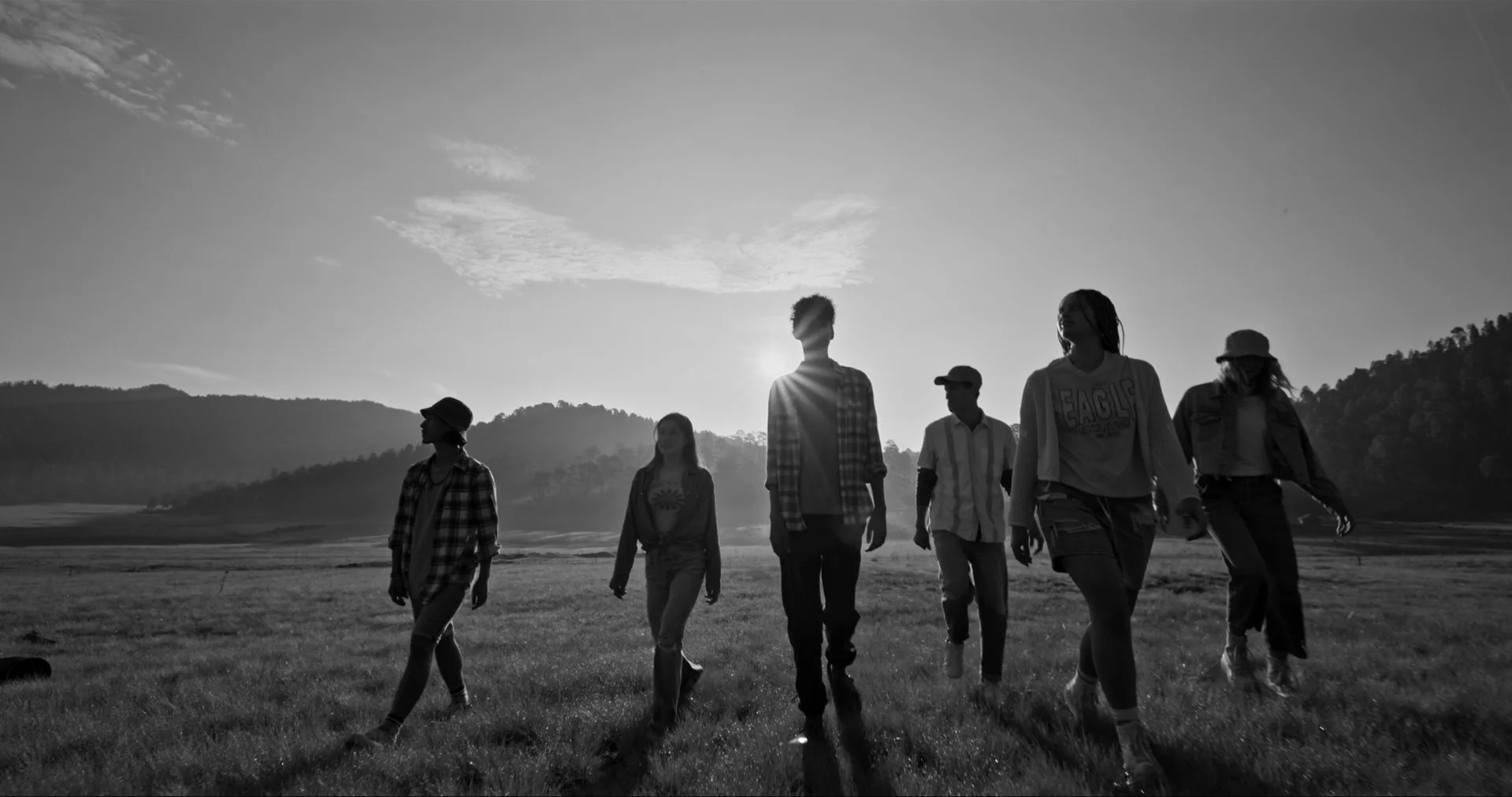 A group of seven young people walking outdoors on a grassy field during sunset, with mountains and clouds in the background, silhouetted against the sky.