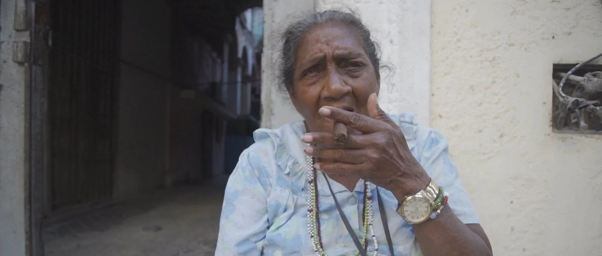 An elderly woman with gray hair and dark skin, wearing a light-colored shirt, multiple necklaces, and a gold wristwatch, touching her lips with a concerned or thoughtful expression, standing outside near a cream-colored wall.