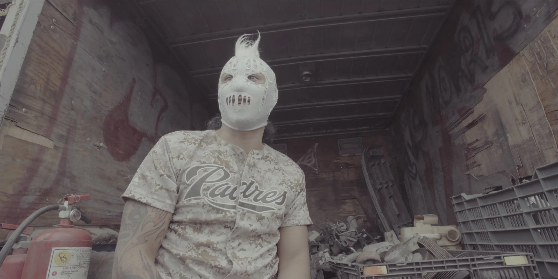 Person wearing a white mask with ghoulish features in a cluttered storage area with automotive parts and debris.