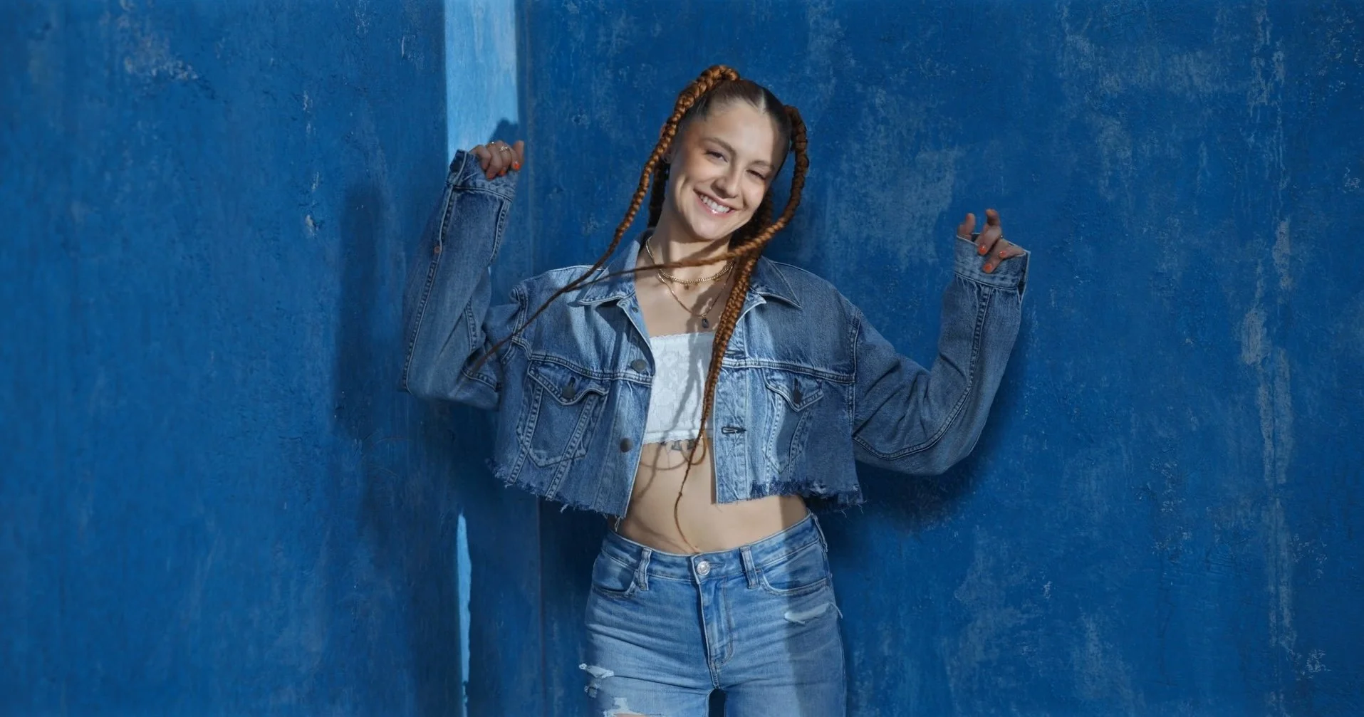 A young woman with braided hair wearing a denim jacket and ripped jeans, smiling and flexing her arms against a blue textured wall.