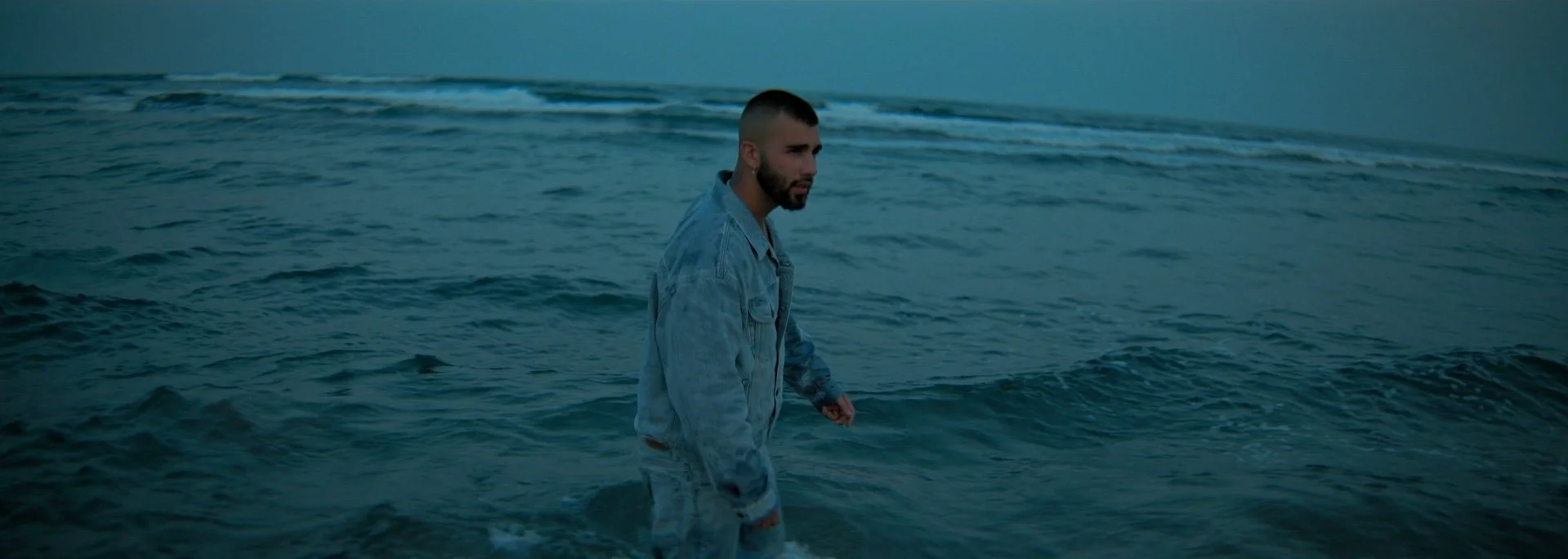 A man with short hair and a beard, wearing a light-colored denim jacket, standing in the ocean near the shore during dusk or dawn.