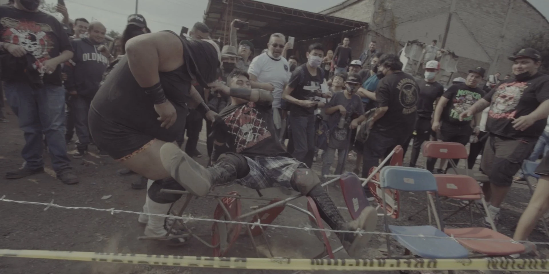 A person dressed in black with a bandana over their face is lying on a bike, while another person stands over them, holding their shirt. A crowd of onlookers surrounds the scene, some taking photos, with chairs and a construction site visible in the