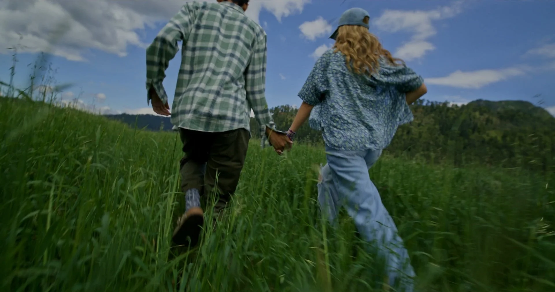 A couple holding hands and walking through a grassy field on a sunny day, with hills and a blue sky with clouds in the background.