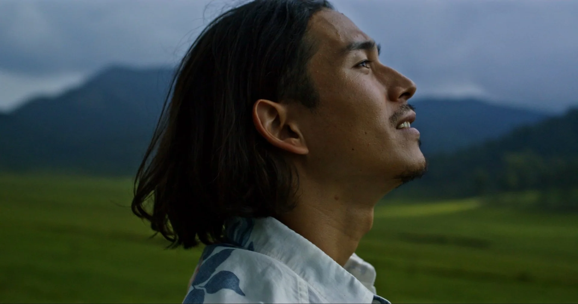 A man with long dark hair and a mustache standing outdoors with mountains and greenery in the background.
