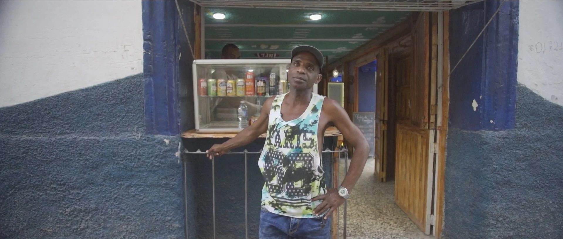 Man in tank top and cap standing in front of a small food stall or shop with wooden doors.