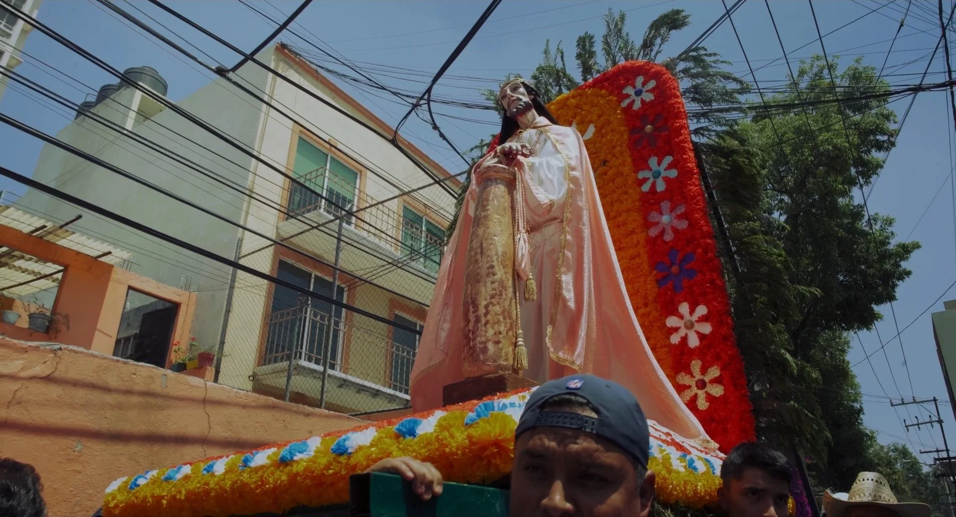 A religious procession float featuring a statue of a saint or religious figure adorned with a flowing pink and white robe, set outdoors with buildings, trees, and power lines in the background. The float is decorated with bright orange and yellow flo