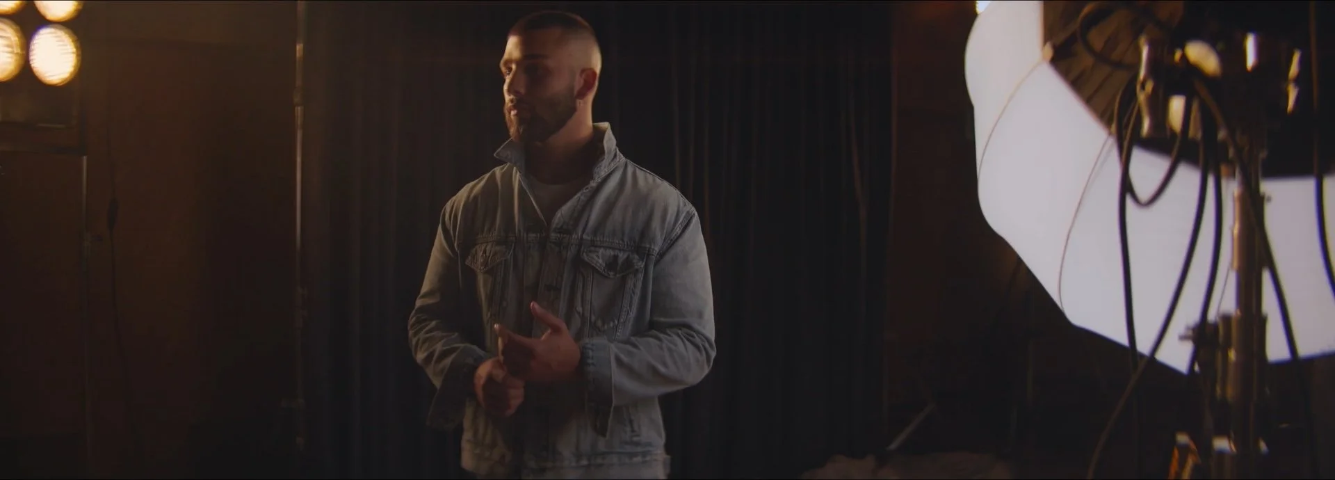 A man with a beard and short hair, wearing a denim jacket, stands in a dimly lit room next to a large professional photography light with a white diffuser. He appears to be in a photography or video studio.