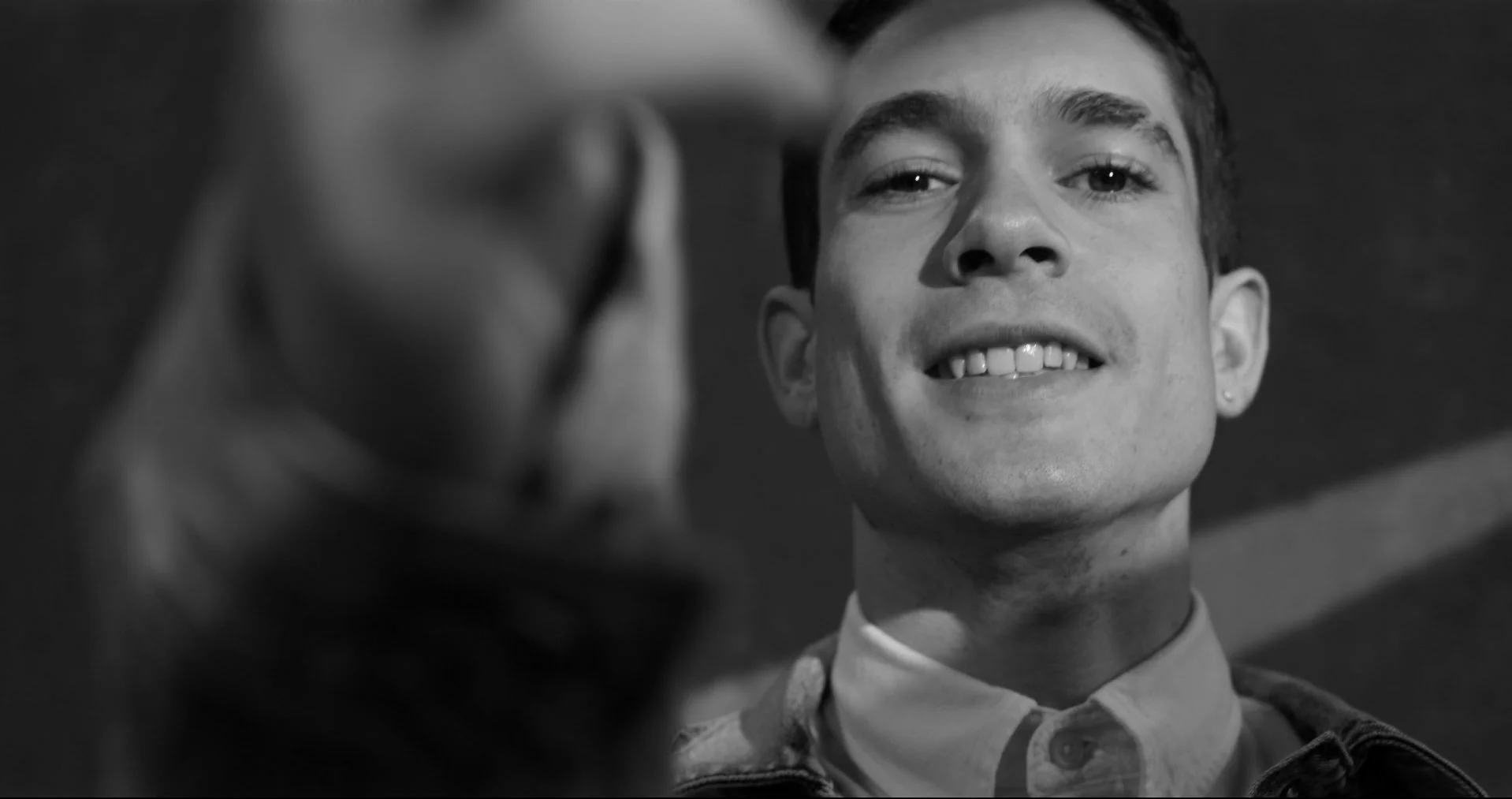 A young man with short dark hair, smiling slightly, in black and white, looking at the camera, with a blurred hand in the foreground.