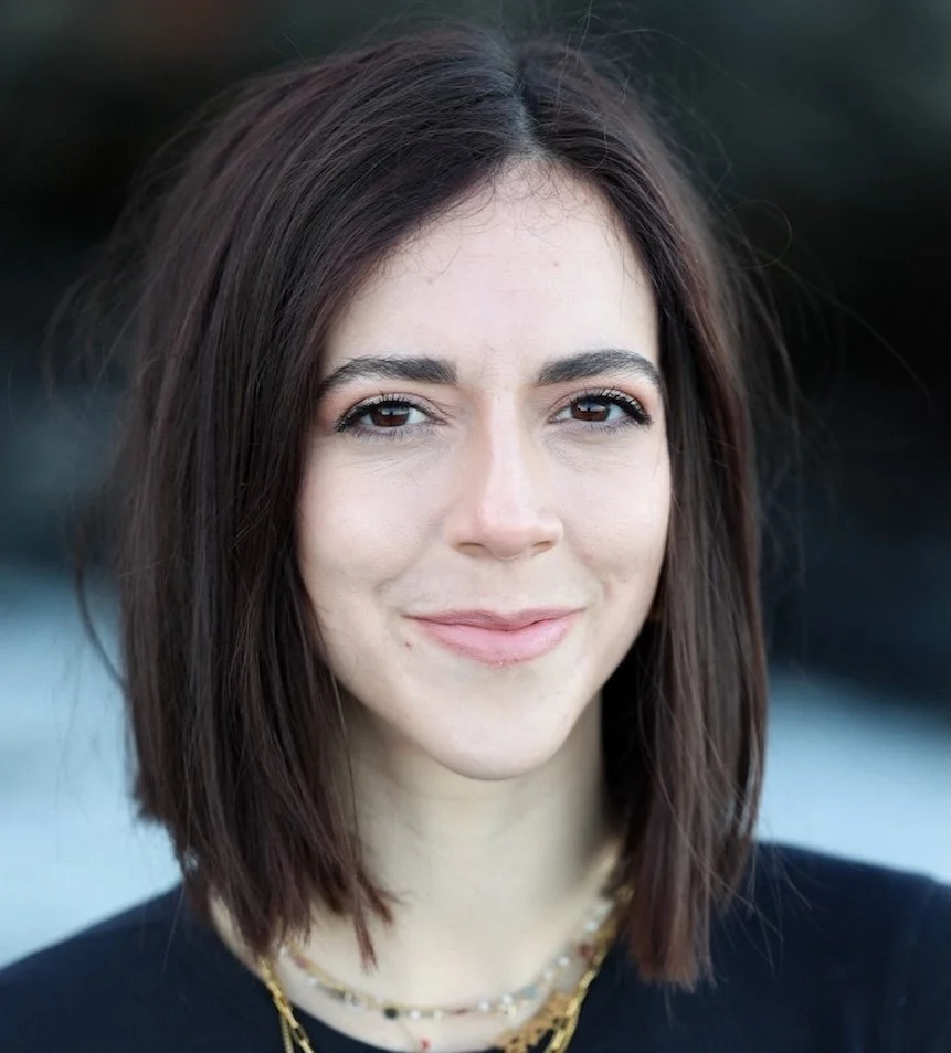 A young woman with shoulder-length dark brown hair, smiling slightly, wearing a black top and layered gold necklaces, outdoors.