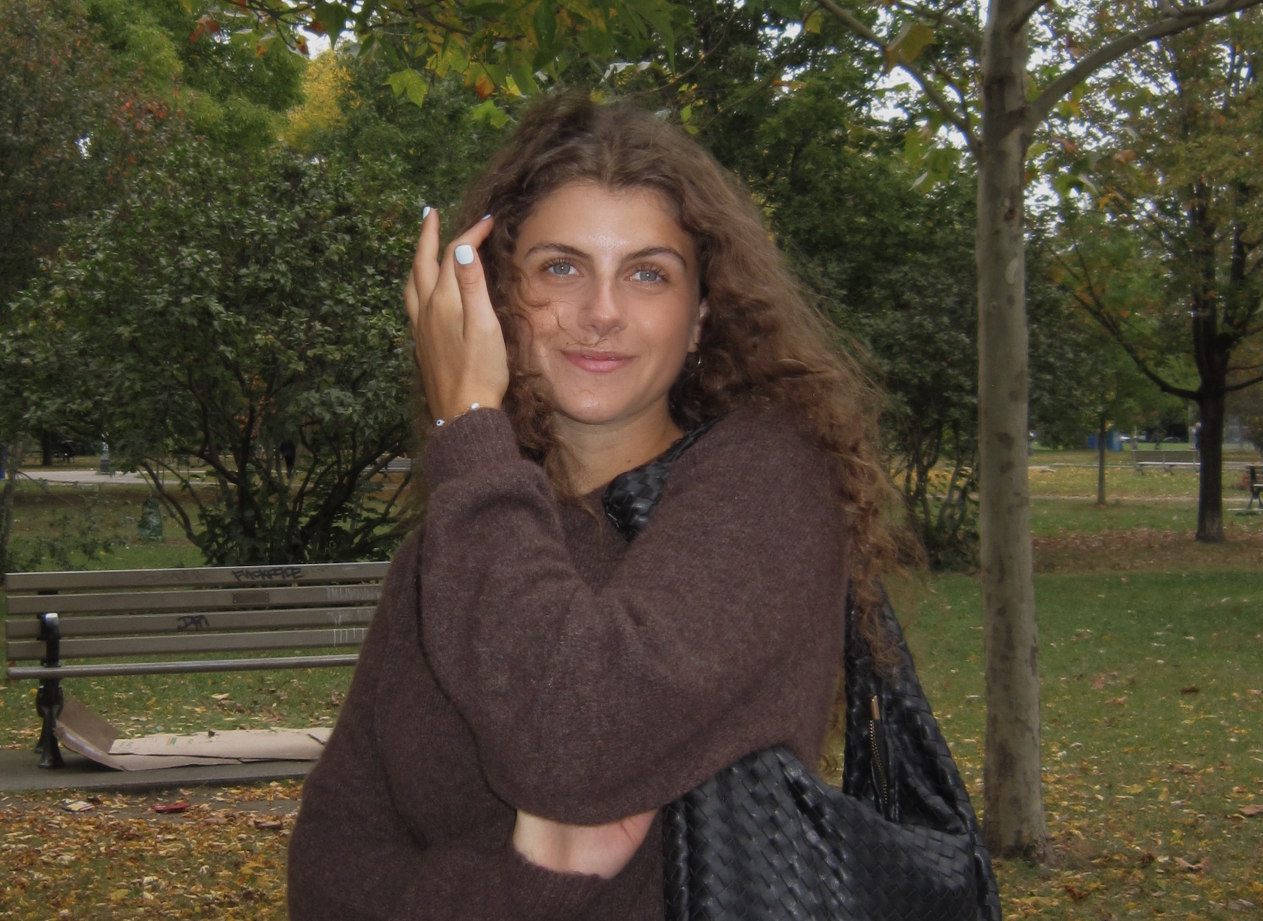 A young woman with long brown hair smiling outdoors, wearing a brown sweater, with a nature background.