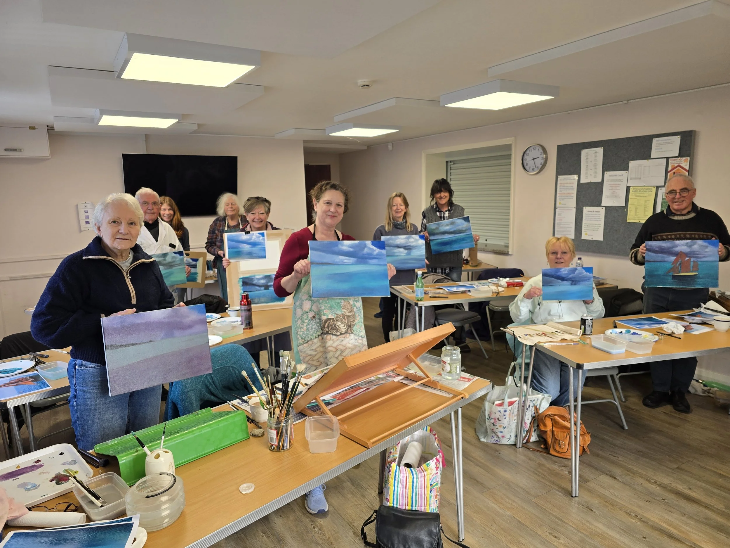 A group of people in an art class holding their completed landscape paintings of the sea with sky, in a well-lit room with tables, art supplies, and a bulletin board.