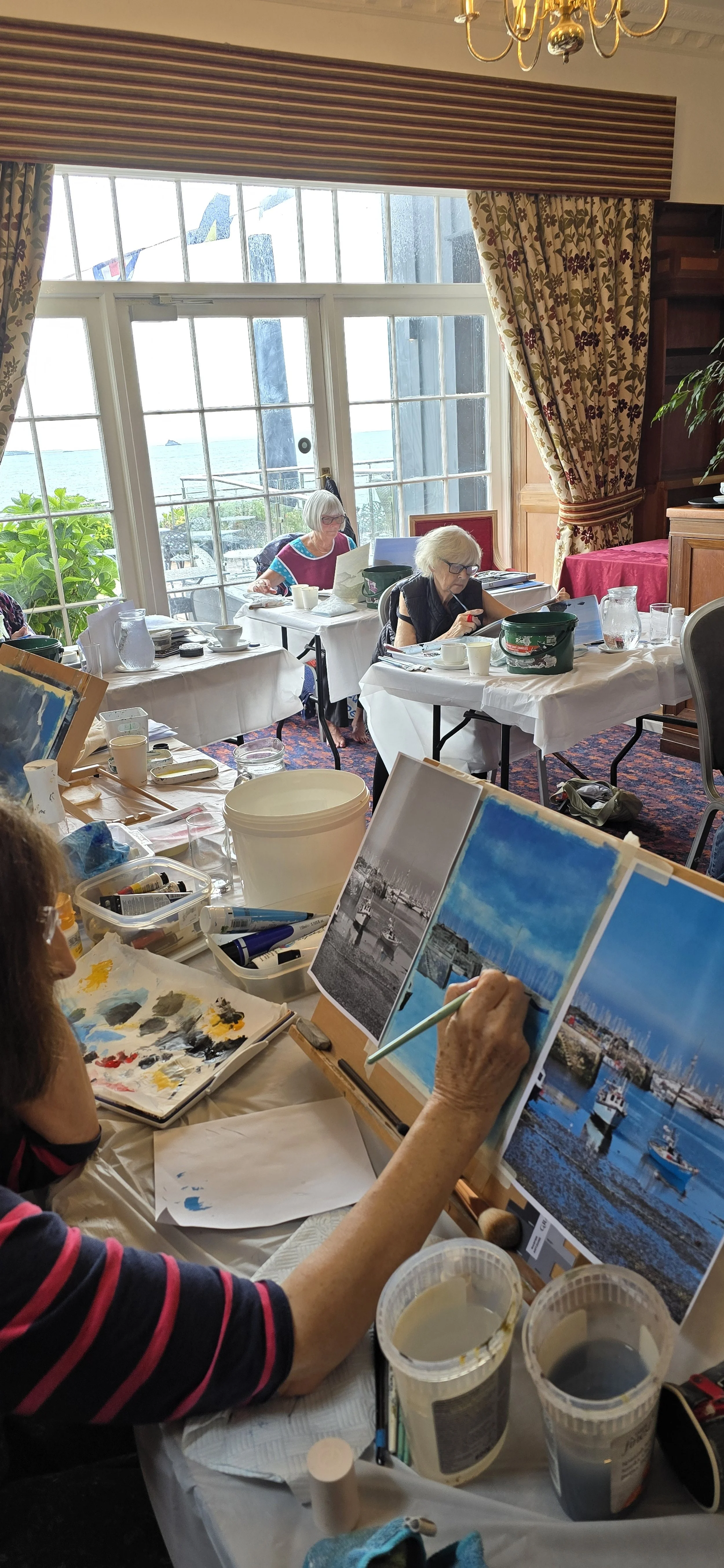 A group of women painting scenic harbor scenes on canvases in a well-lit room with large windows and sea view.