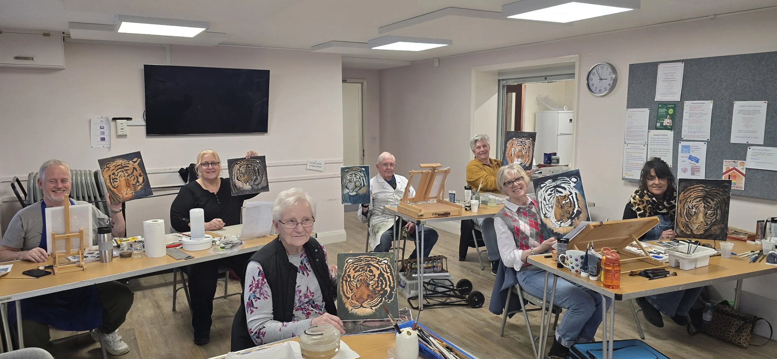 Group of seven diverse adults in a classroom, each holding a tiger painting they created during a painting class.
