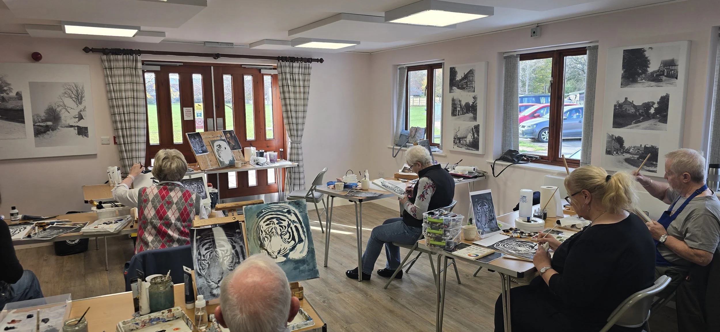 Adults painting black and white portraits of tigers and landscapes in an art class room with windows showing parked cars outside.