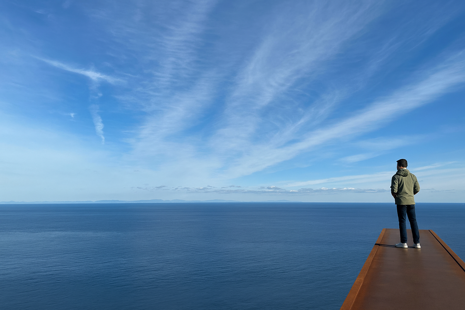 A man in a khaki green hooded jacket, dark blue jeans, and white sneakers stands on a long, rusted iron beam cantilevered over a steep slope; the man is looking over the clear blue water of Tokyo Bay and a blue sky with streaks of white clouds