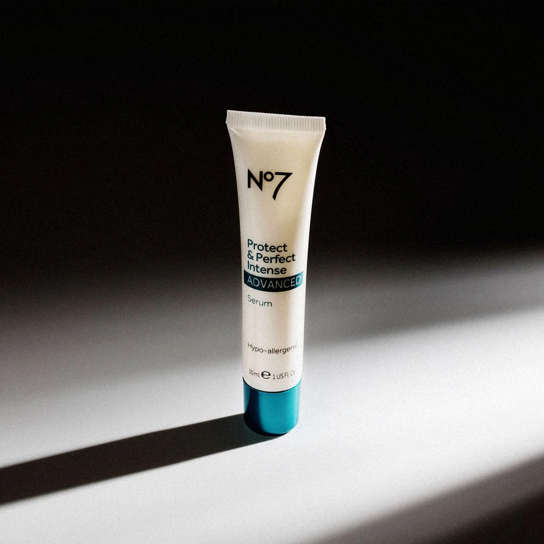A tube of No7 Protect & Perfect Intense Advanced Serum standing upright on a surface with a shadow cast to the left, against a black and white gradient background.