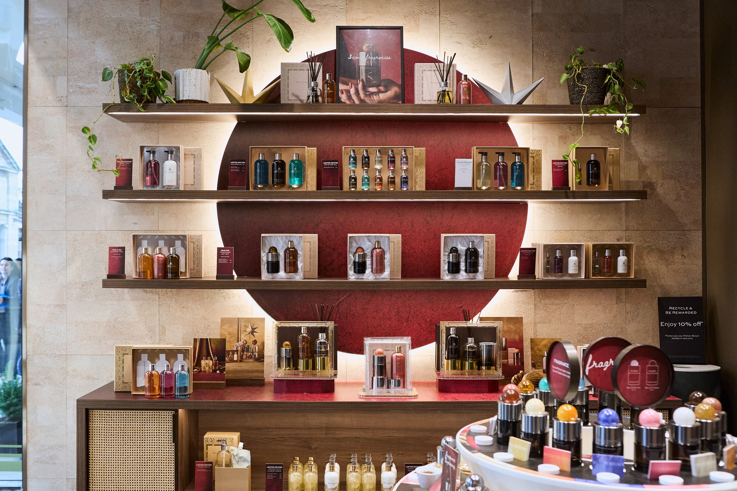 Display of perfume bottles arranged on shelves and a table in a retail store, with decorative plants and promotional signs.