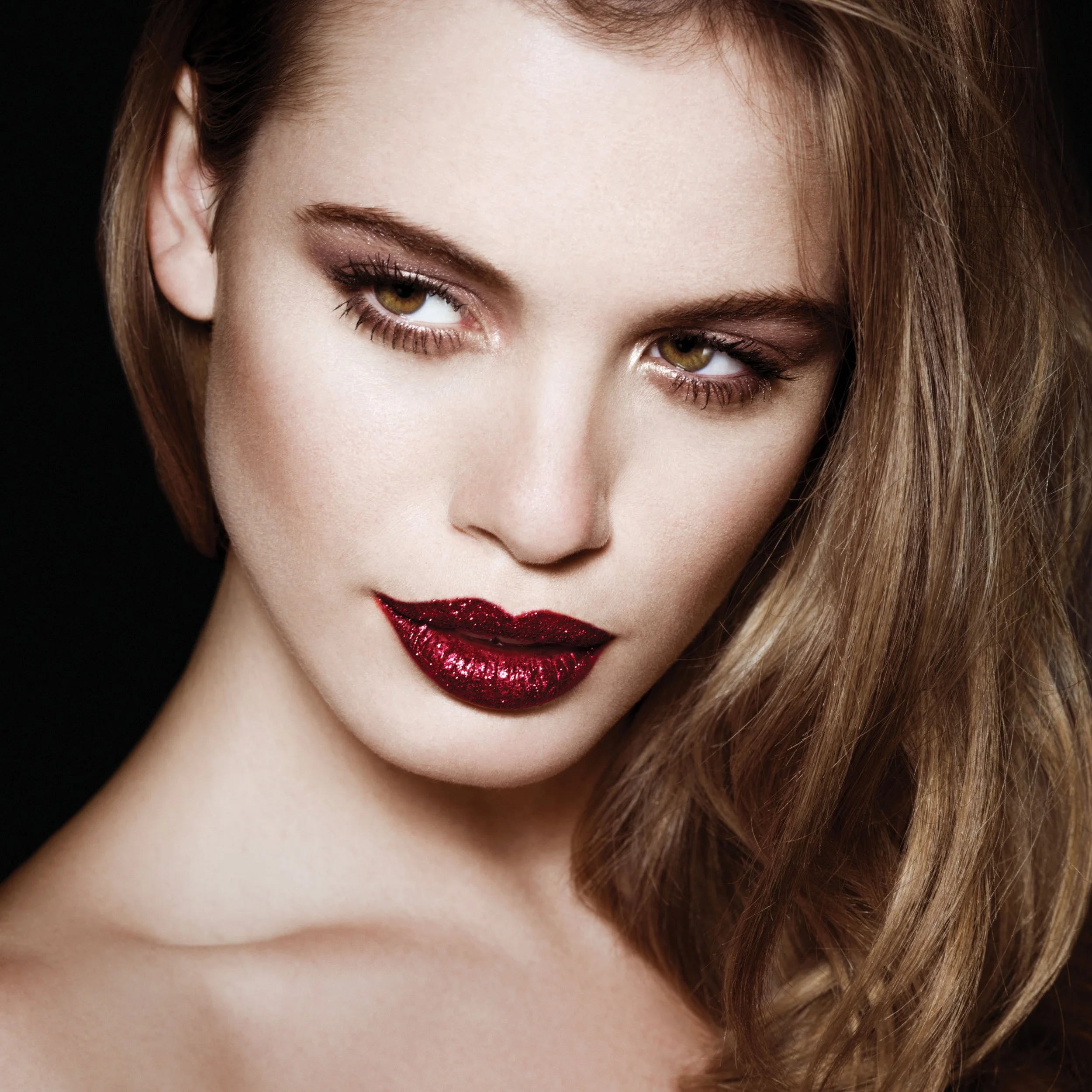 A woman with fair skin, brown hair, and hazel eyes having makeup with dark lipstick and eye shadows, looking to the side with a slight smile.