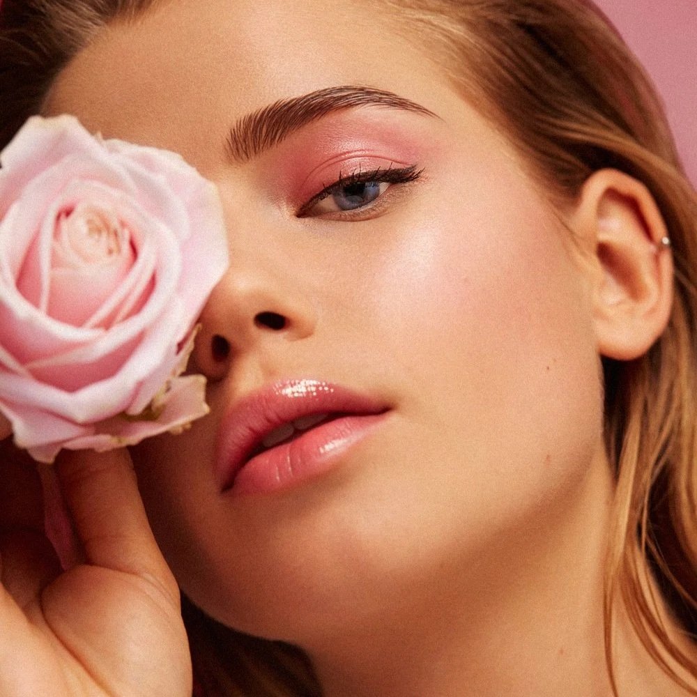 Close-up of a woman holding a pink rose close to her face with pink makeup and glossy lips.