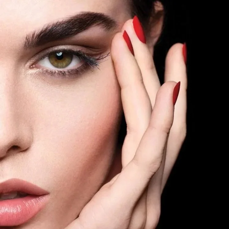 Close-up of a woman with light skin, green eye, and makeup, touching her face with fingers that have red nail polish.
