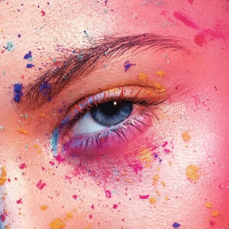 Close-up of a person's eye with blue iris, decorated with colorful birthday or festival makeup and glitter in pink, blue, and gold.