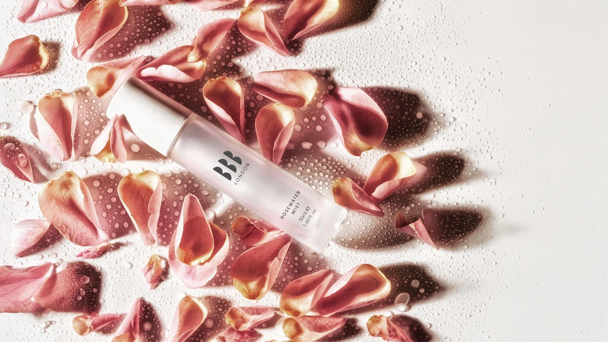 A bottle of BBB London Rosewater Mist surrounded by rose petals and water droplets on a white surface.