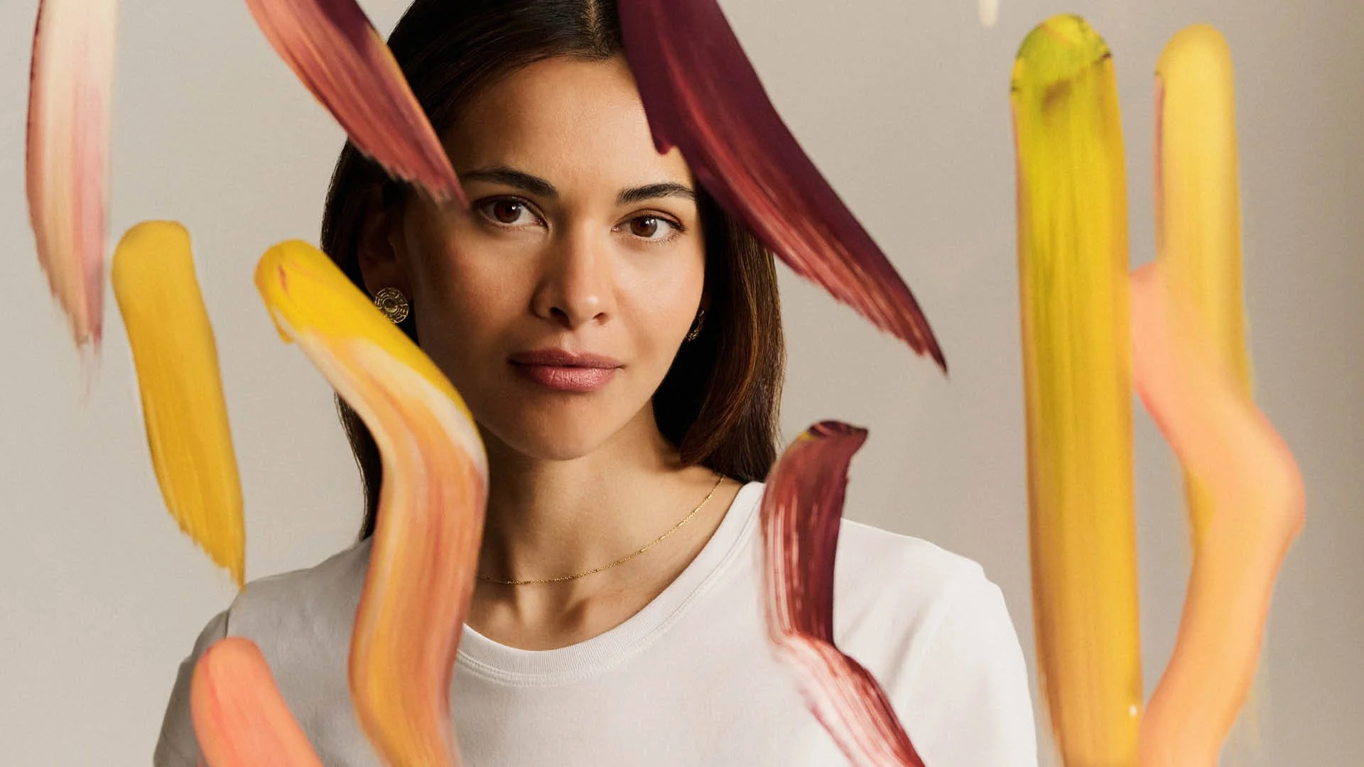 A woman with dark hair and light skin looking through several colorful brushstrokes, with a neutral background, wearing a white top, gold earrings, and a delicate gold necklace.