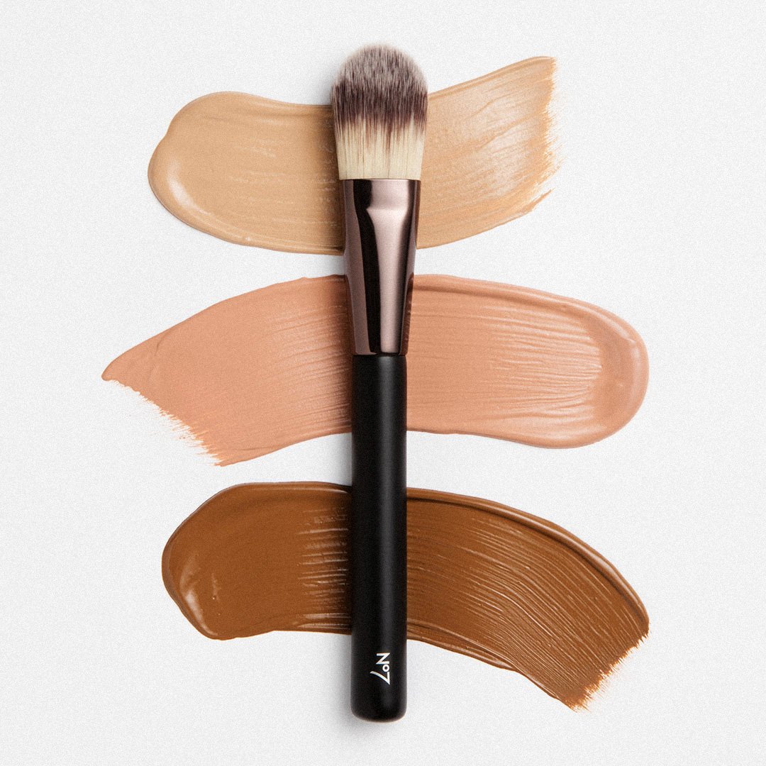 A makeup brush with beige, peach, and brown foundation swatches behind it on a white background.