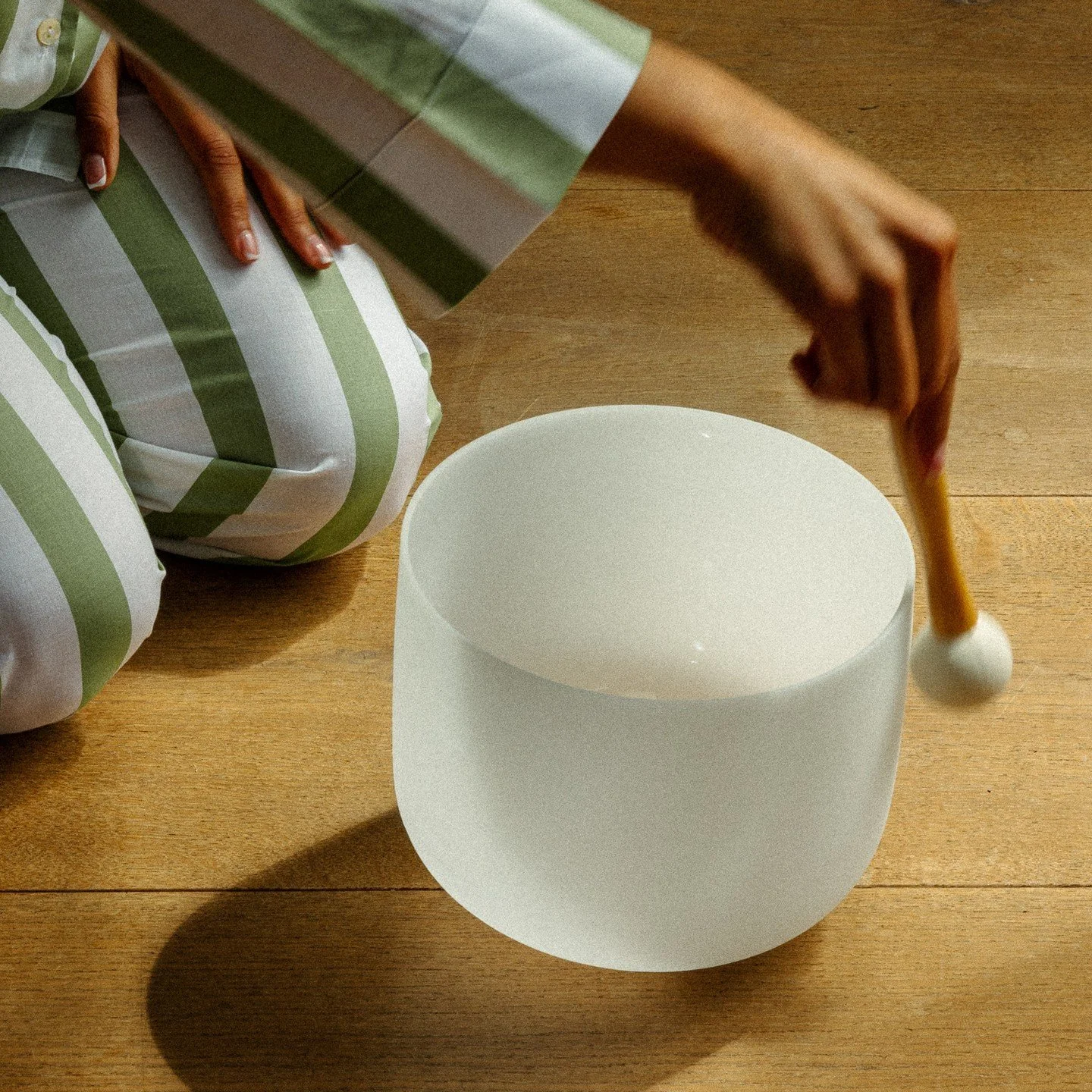 A person wearing striped green and white pants plays a white singing bowl with a wooden hammer.