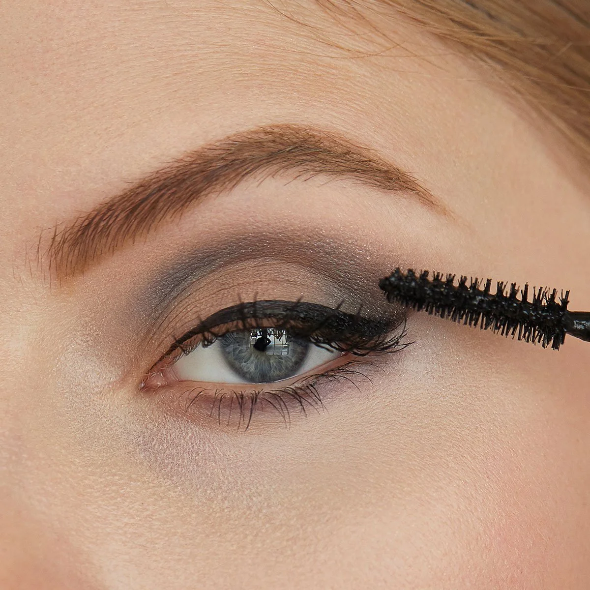 Close-up of a woman applying black mascara to her upper eyelashes with a wand, showcasing eye makeup with smoky eyeshadow, well-groomed eyebrows, and blue eyes.