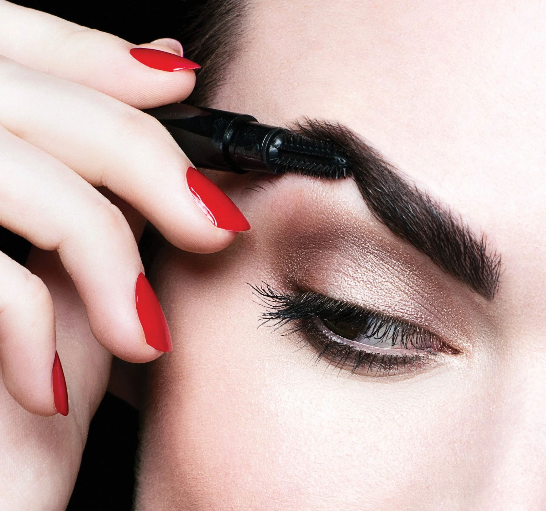 Close-up of a woman applying makeup using a black eyebrow brush, with red nail polish visible on her fingers.