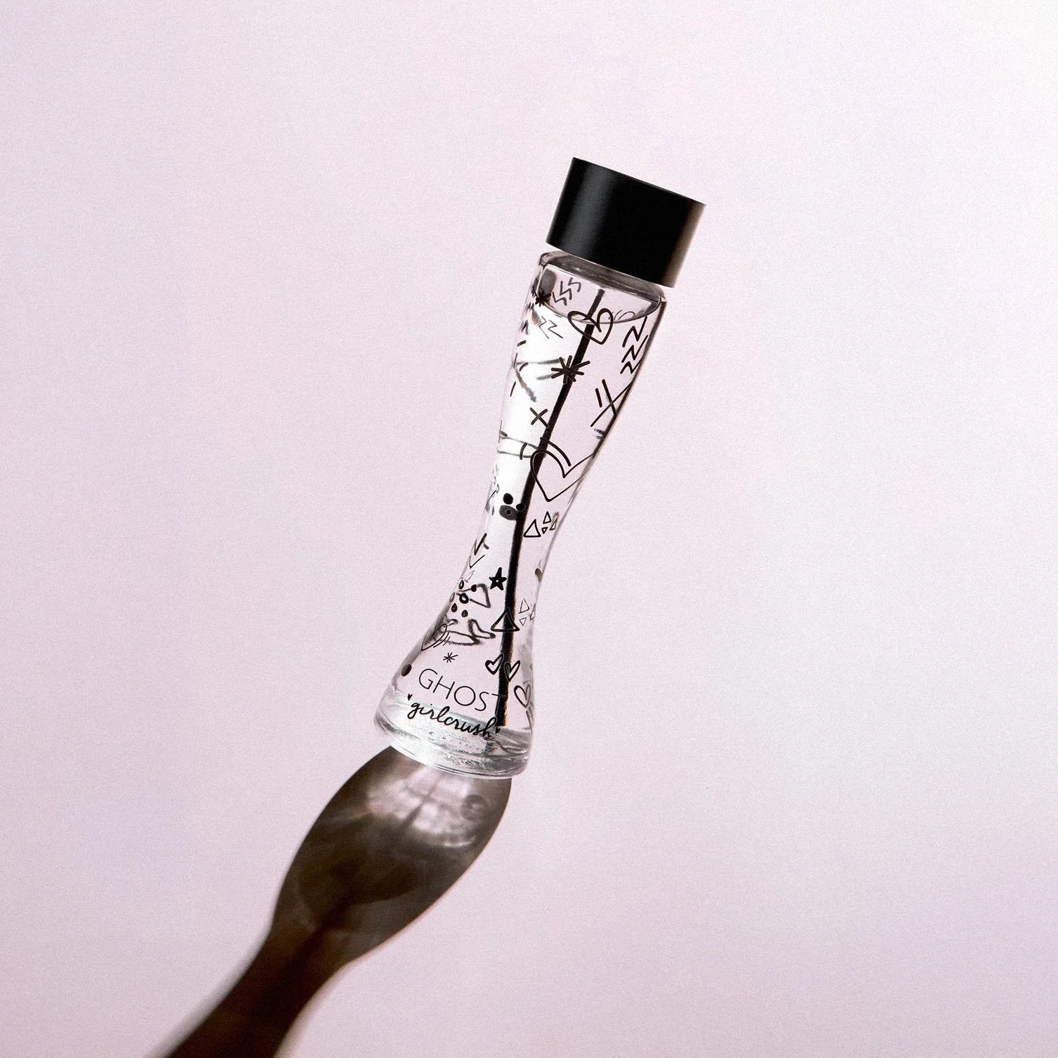 A clear glass perfume bottle with black cap, decorated with black doodles and icons, casting a shadow on a plain light background.