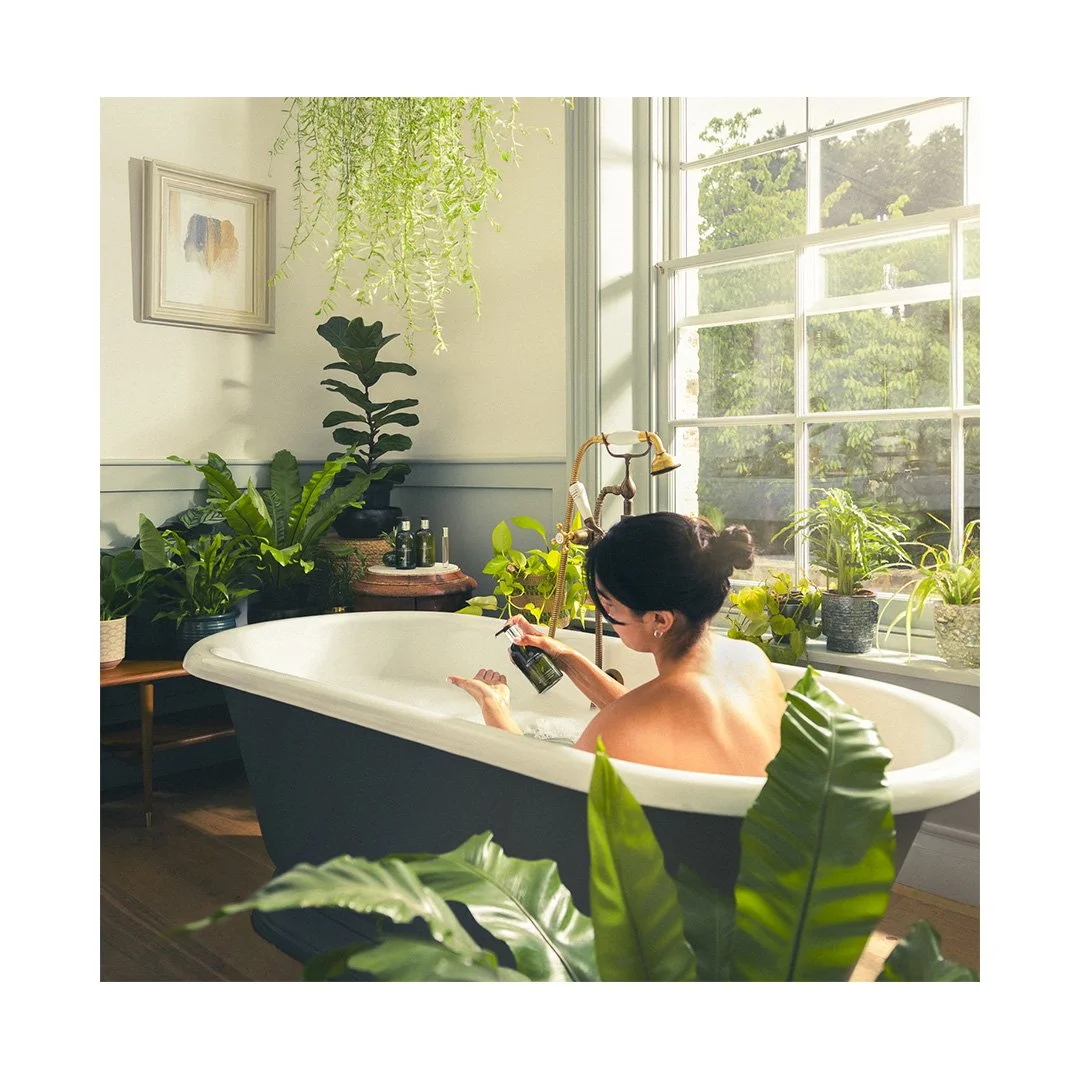A woman taking a bath in a vintage bathtub surrounded by green plants in a bright, sunlit room with large windows.