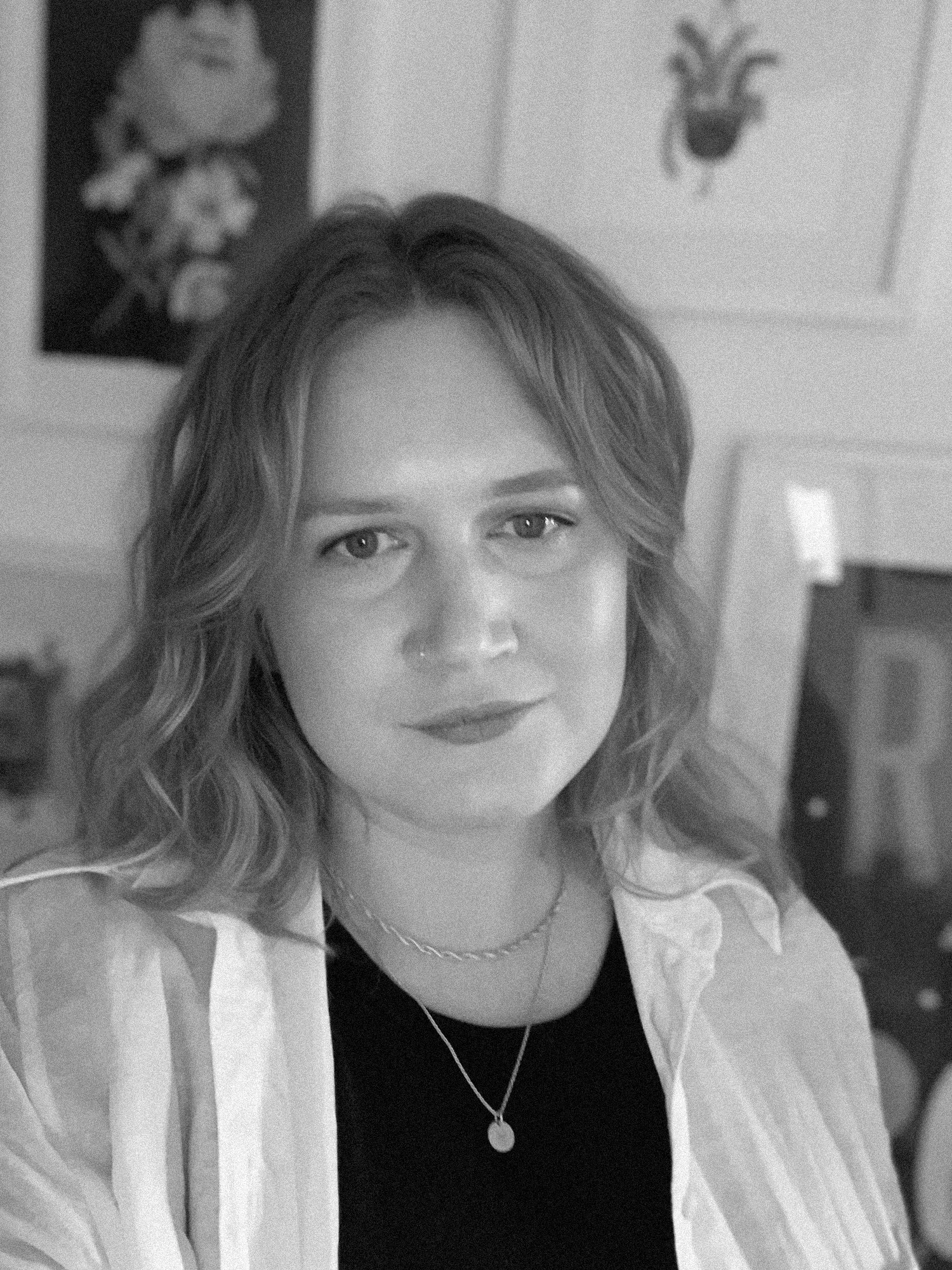 A woman with wavy hair, wearing a light-colored striped shirt and layered necklaces, looking at the camera. Artwork hanging on the wall behind her.