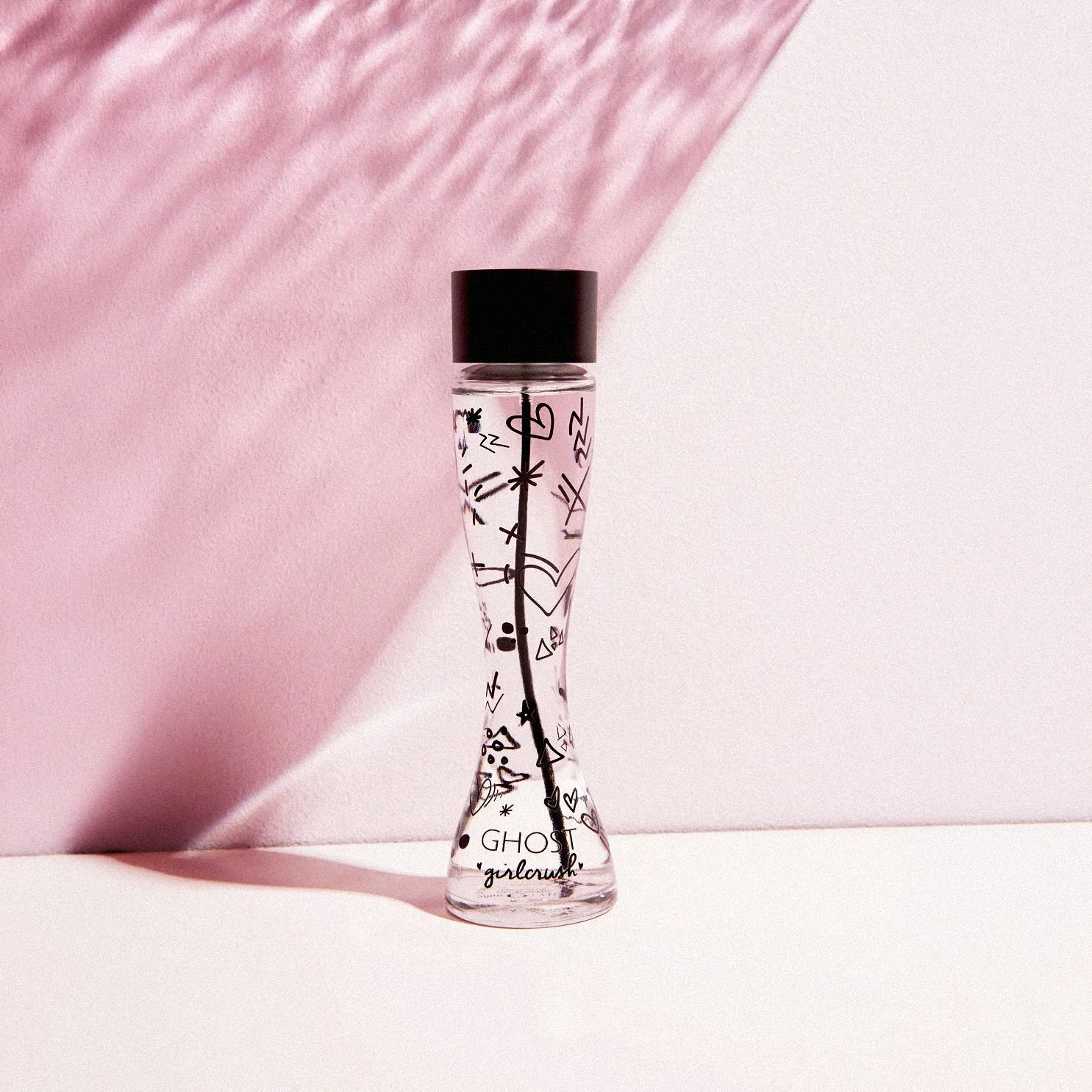 A clear glass bottle with black doodle-style drawings and the text 'GHOST girl crush' on it, standing against a pink and white background.