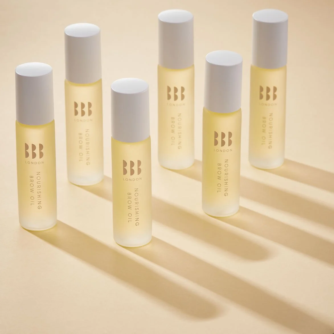 Six bottles of nourishing brow oil with white caps and transparent yellowish liquid, arranged on a light surface casting shadows.