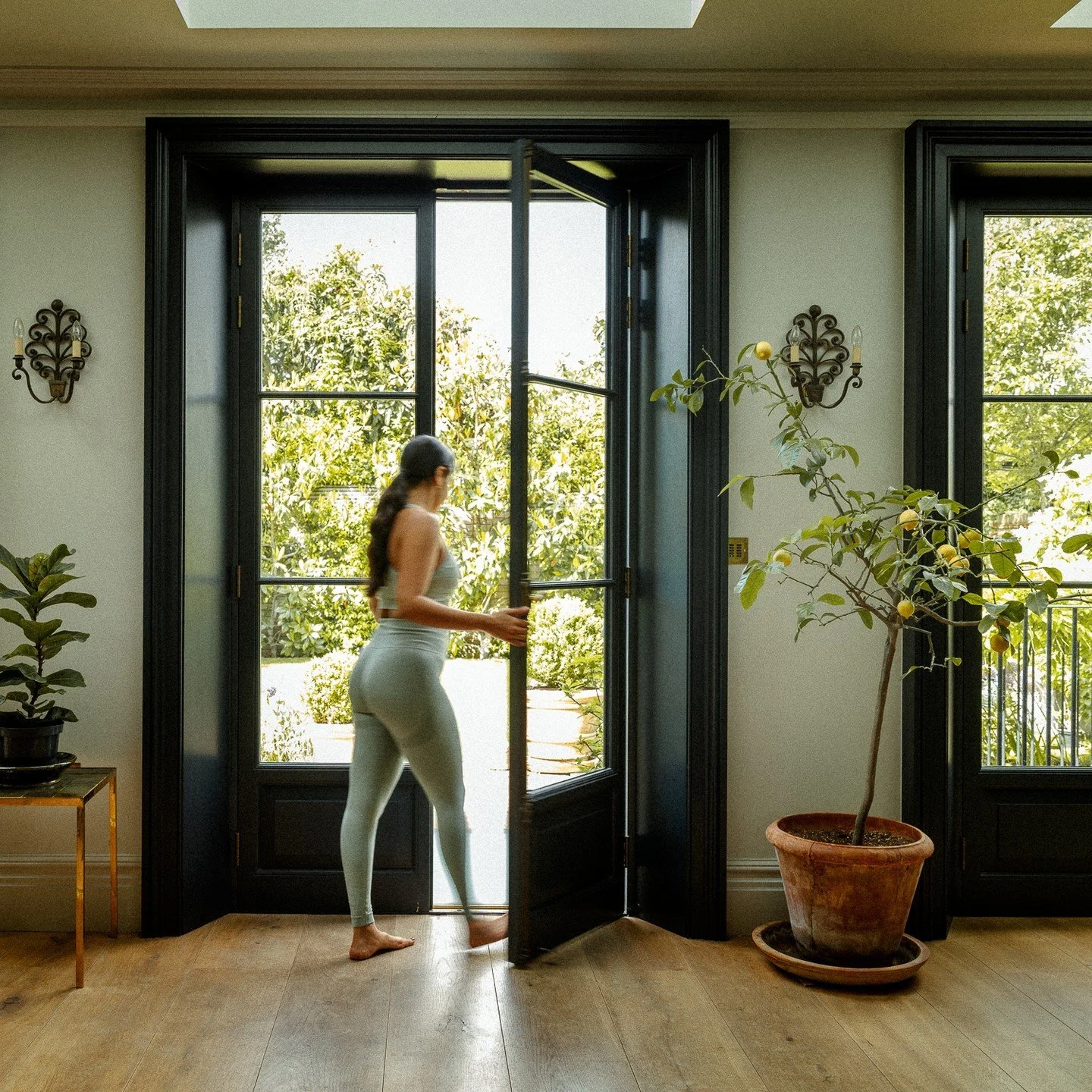 A woman in athletic wear opens a door to a sunlit garden with trees and plants inside a room with black window frames and white walls, decorated with potted plants and wall sconces.