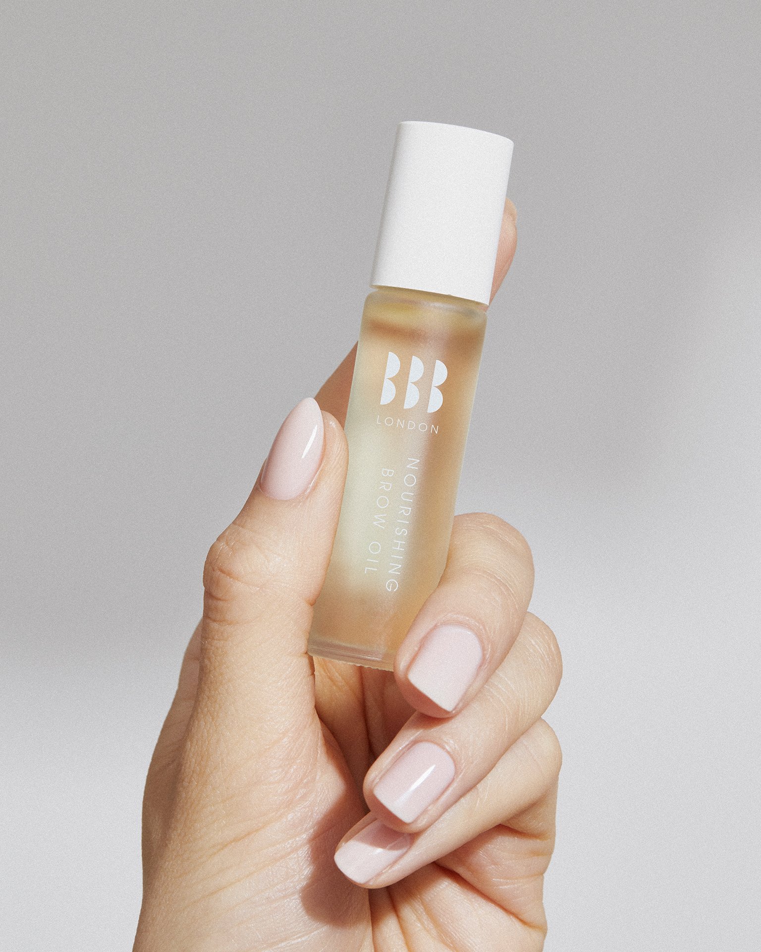 A hand holding a small frosted glass bottle of BB London nourishing brow oil with an off-white cap, against a light gray background.