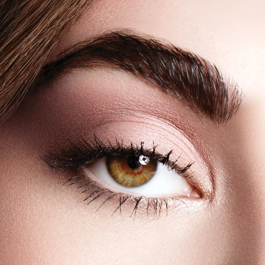 Close-up of a woman's eye with brown iris, well-groomed eyebrow, and makeup including pink eyeshadow and mascara.