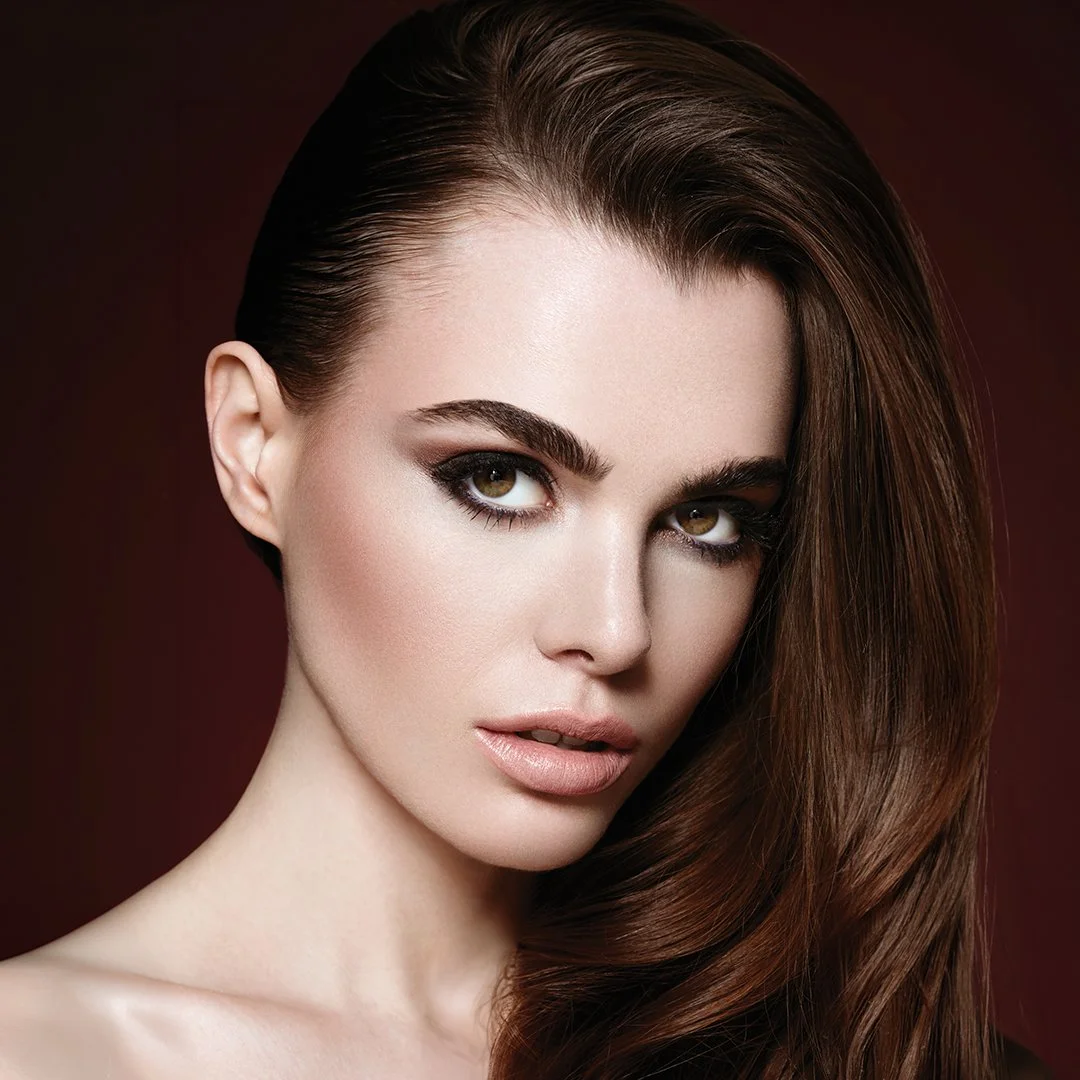 Close-up portrait of a woman with brown hair and makeup, against a dark red background.