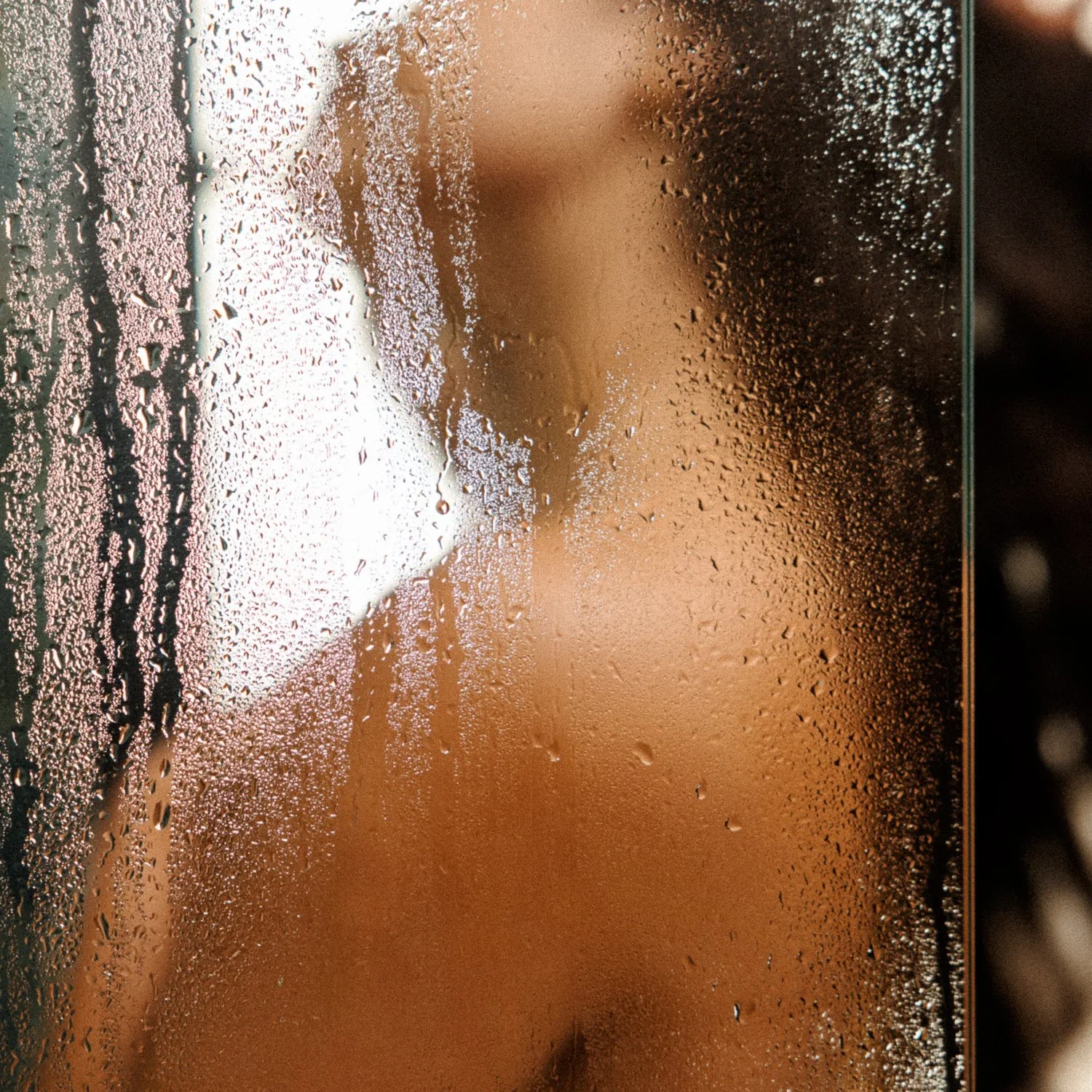 A person with long hair behind a foggy glass shower door with condensation and water droplets.