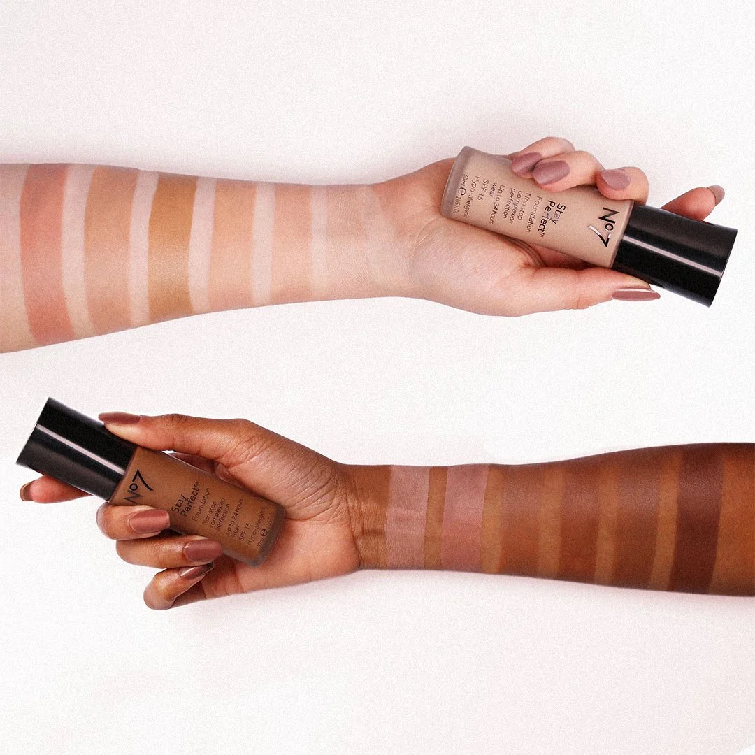 Two arms with various shades of foundation swatches and two bottles of foundation held in hands.