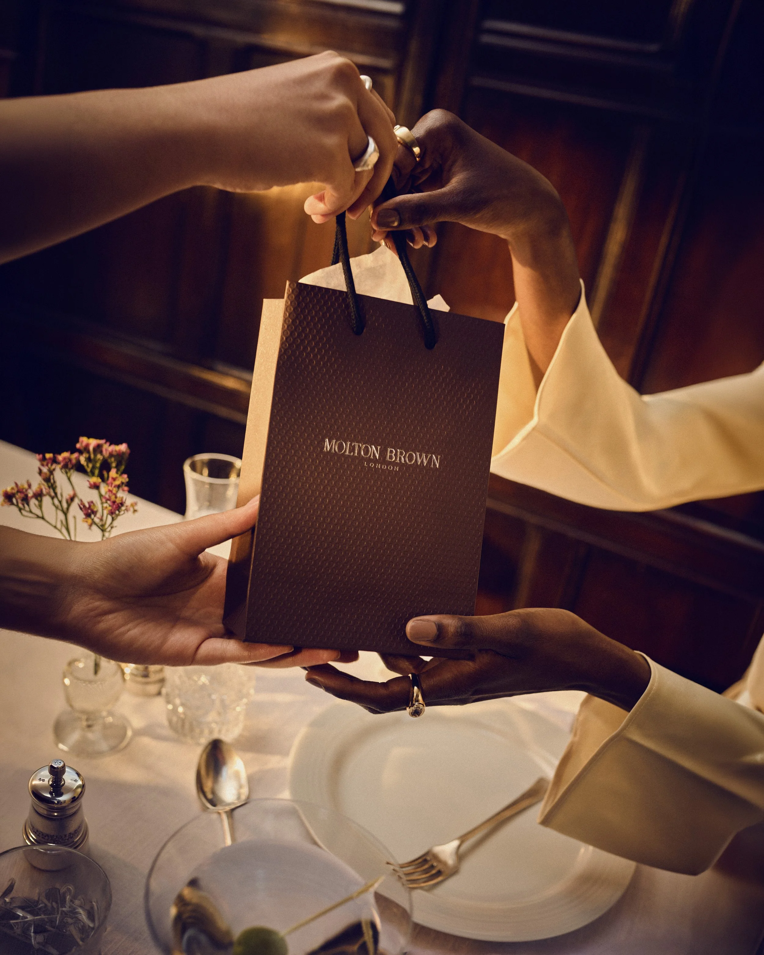 Two people exchanging a Molton Brown London gift bag at a formal event, with tableware and flowers visible on the table.
