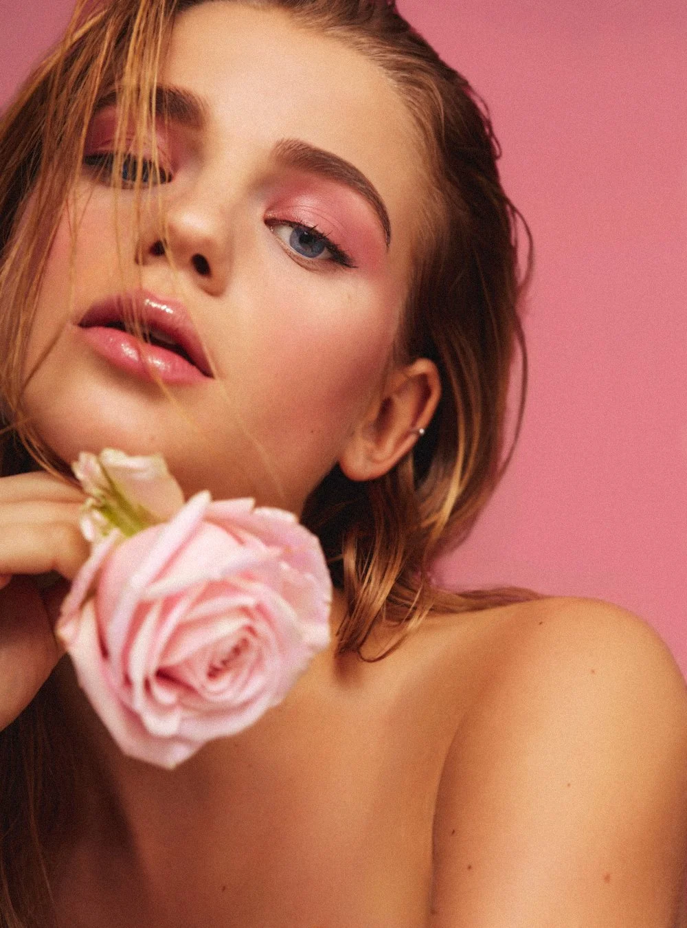 Close-up of a young woman with curly red hair and pink eye makeup, holding a pink rose against a pink background.