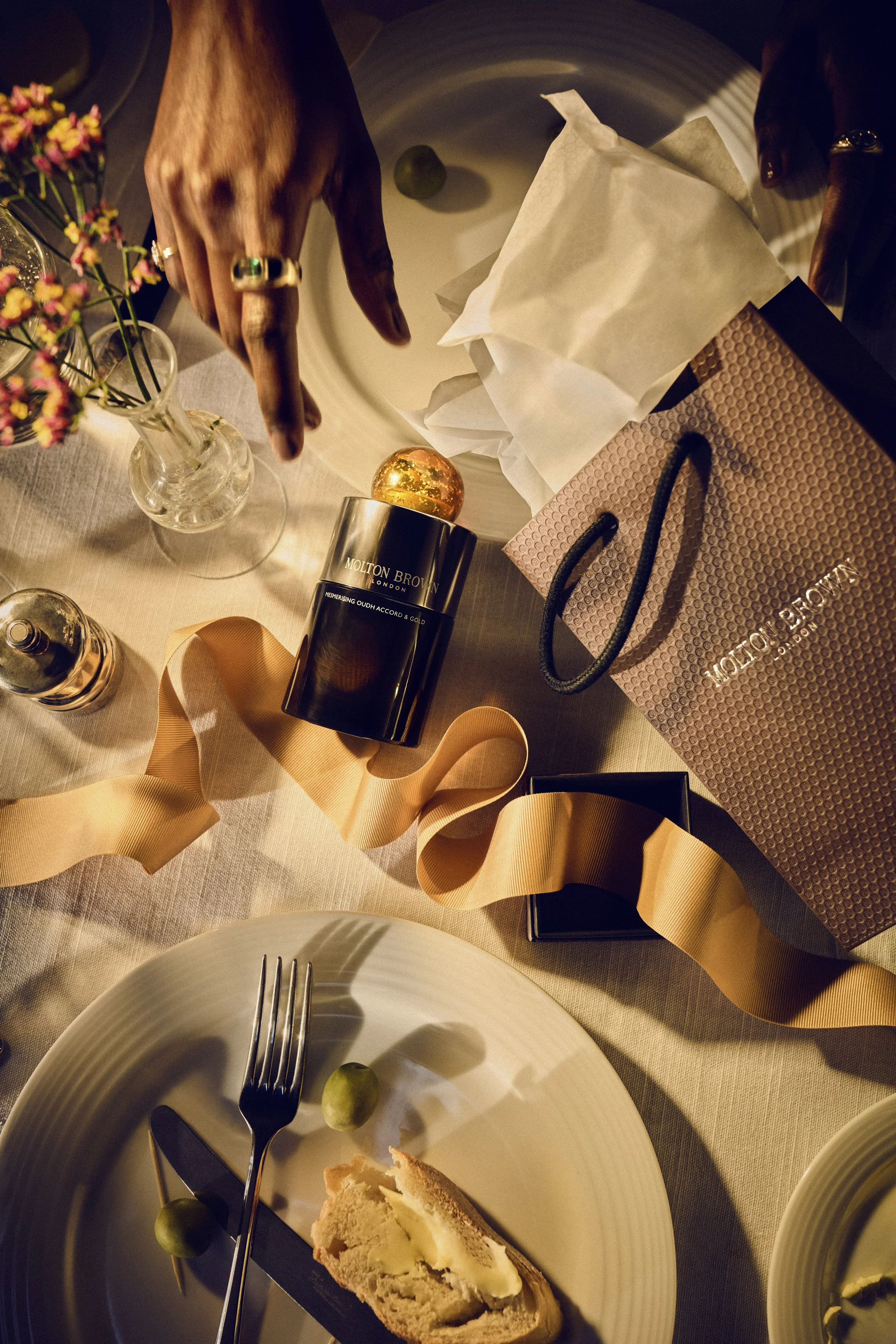 A table setting with a white plate holding a slice of bread with butter, a fork, and a green olive. There is a small vase with pink and yellow flowers, a gold and black Molton Brown skincare product, and a pink shopping bag on the table. The image is warmly lit and slightly tilted.
