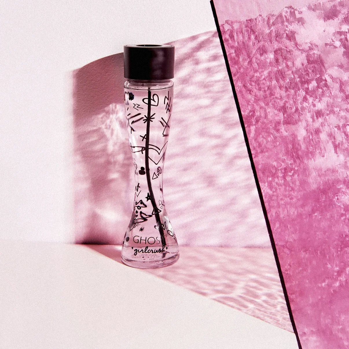 A clear glass perfume bottle with black doodles and words, set against pink textured backgrounds with shadows.