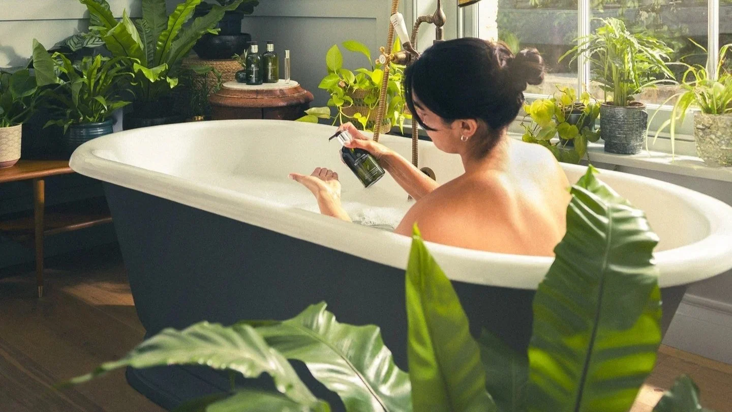 Woman with dark hair in a bun relaxing in a white bathtub filled with bubbles, surrounded by green potted plants, and holding a bottle of bath product while sitting near a window.