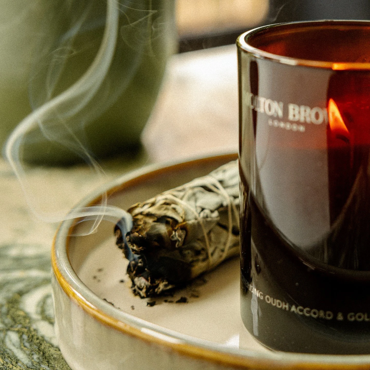A burning sage smudge stick next to a black candle in a black container with white text on a white and gold-edged plate.