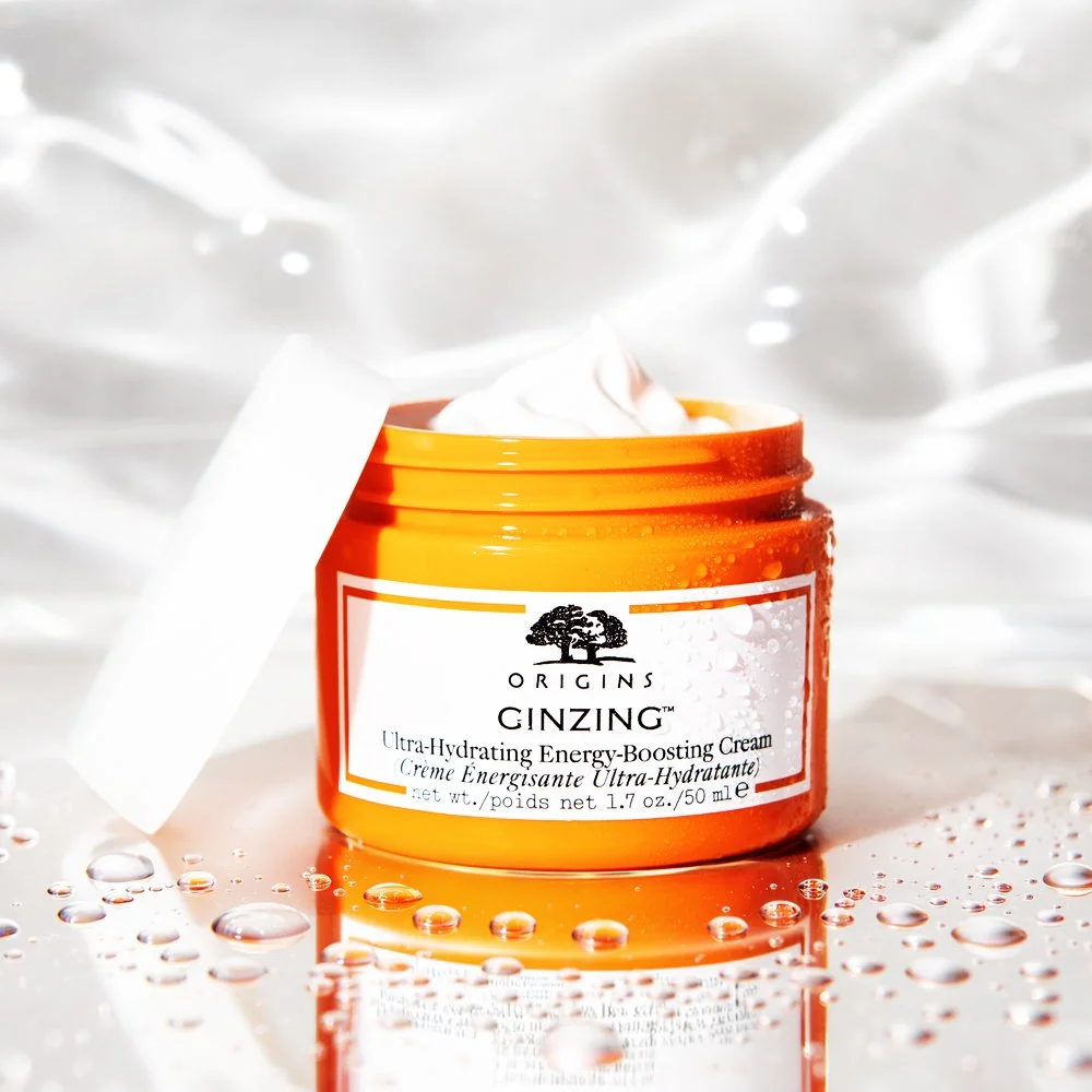 Open jar of Origins Ginzing Ultra-Hydrating Energy-Boosting Cream with cream visible inside, on a reflective surface with water droplets and a blurred light background.