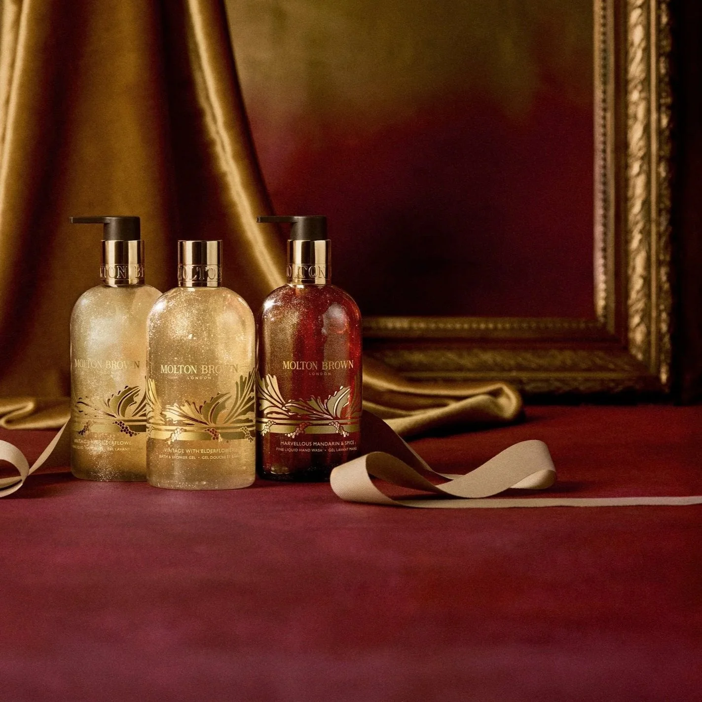 Three bottles of Molton Brown hand wash and gel, with two bottles of golden-colored products and one bottle of red-colored product, set against a gold satin background and a framed mirror.